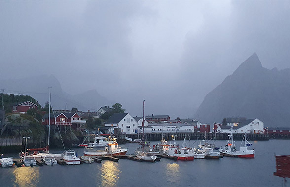 La nebbia scende sulla città di Hamnøy, in Norvegia. Le luci delle case si riflettono sull'acqua del porto, dove diverse imbarcazioni sono ormeggiate.